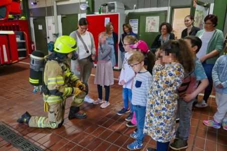 Bild von einer Gruppe von Schüler*innen. Vor den Schüler*innen kniet ein Feuerwehrmann, der eine Atemschutzmaske im Gesicht und eine Sauerstoffflasche auf dem Rücken trägt.