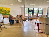 Bild vom Wahlbüro. An einem Tisch sitzt der 4-köpfige Wahlvorstand. In 2 Wahlurnen geben Schüler*innen ihre Stimme ab.