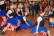 Bild von einer Gruppe von Mädchen, die alle das gleiche Kostüm tragen und tanzen. Rundherum stehen Zuschauer*innen die in die Hände klatschen.