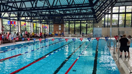Bild von einem Schwimmbecken in einem Hallenbad. Die einzelnen Bahnen sind abgetrennt. Am Rand sind viele Zuschauer*innen.