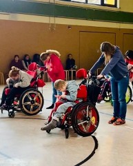 Bild von 2 Menschen im Rollstuhl. Hinter ihnen stehen 2 Begleiterinnen. Alle 4 Personen beugen sich nach vorne und verharren in der Position.