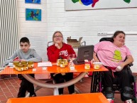 Bild von 3 Sch&uuml;ler*innen, die hinter einem Tisch sitzen. Auf dem Tisch stehen verschieden gro&szlig;e Gl&auml;ser Honig, Probierst&auml;bchen, und T&uuml;ten mit Kerzen.