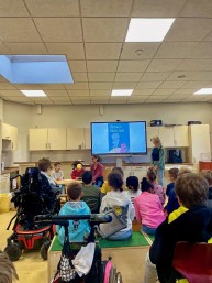 Bild von einer Vorlese-Veranstaltung. im Vordergrund sitzen Sch&uuml;ler*innen im Publikum. Im Hintergrund sitzt zwei Sch&uuml;ler*innen an einem Tisch. Auf einem Fernseher ist das Cover eines Buchs zu sehen.