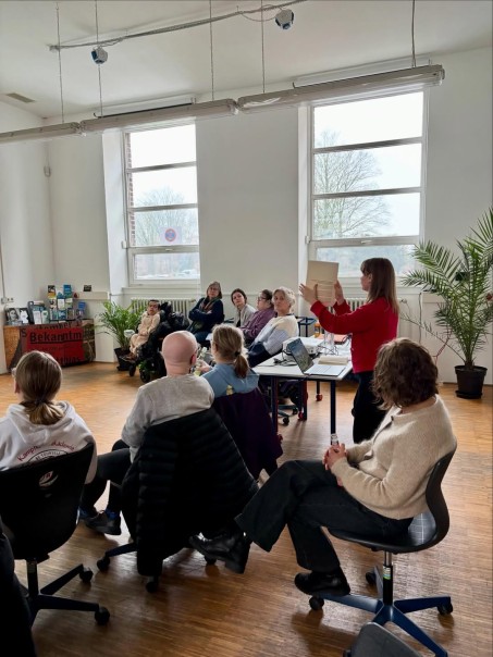 Gruppe von Personen sitzt im Halbkreis. Eine Person h&auml;lt ein Dokument hoch und spricht zu der Gruppe.
