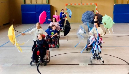 Bild von 8 Sch&uuml;ler*innen im Rollstuhl. Die Betreuer*innen halten einen Regeschirm und schieben die Sch&uuml;ler*innen durch die Turnhalle.