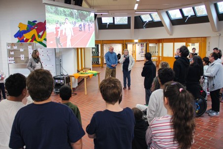Bild von vielen Schüler*innen, die auf eine Leinwand schauen. Vor der Leinwand steht ein Mann und eine frau, die zu den Schüler*innen sprechen.